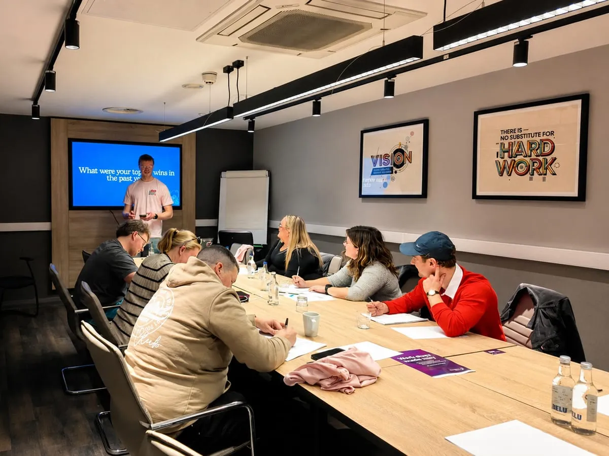 Nathan presenting at the head of a boardroom table while six members write in their workbooks