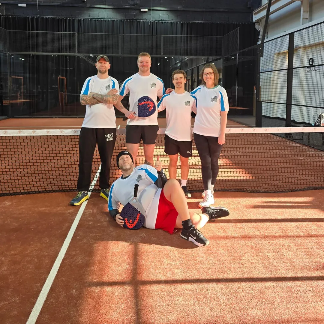 Five CBK members in matching shirts on a padel court, one lying across the front giving a thumbs up